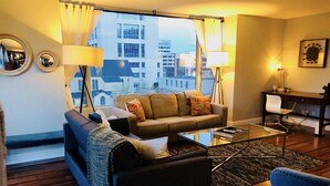 Ein modernes Wohnzimmer mit Sofa, Sessel und Couchtisch. Durch große Fenster ist ein Blick auf Gebäude möglich.