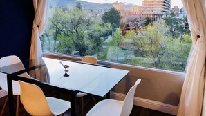 Ein modernes Zimmer mit Glastisch und weißen Stühlen, einem großen Fenster mit Vorhängen sowie Blick auf Stadt und Bäume.