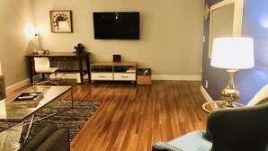 Ein Wohnzimmer mit Flachbildfernseher, Schreibtisch, Stuhl, Sofa und Lampe.