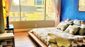 Ein Schlafzimmer mit großem Fenster, einem Bett mit Holzrahmen, einer Bank und einem Schreibtisch.