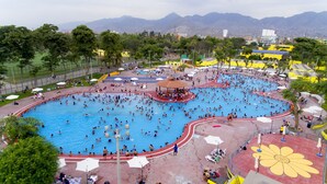 Una gran piscina al aire libre con varias pistas de natación, rodeada de árboles y un paisaje urbano de fondo.