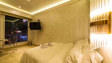 A hotel room with a bed, a TV mounted on the wall, a window with a view of the city at night, and a chair.