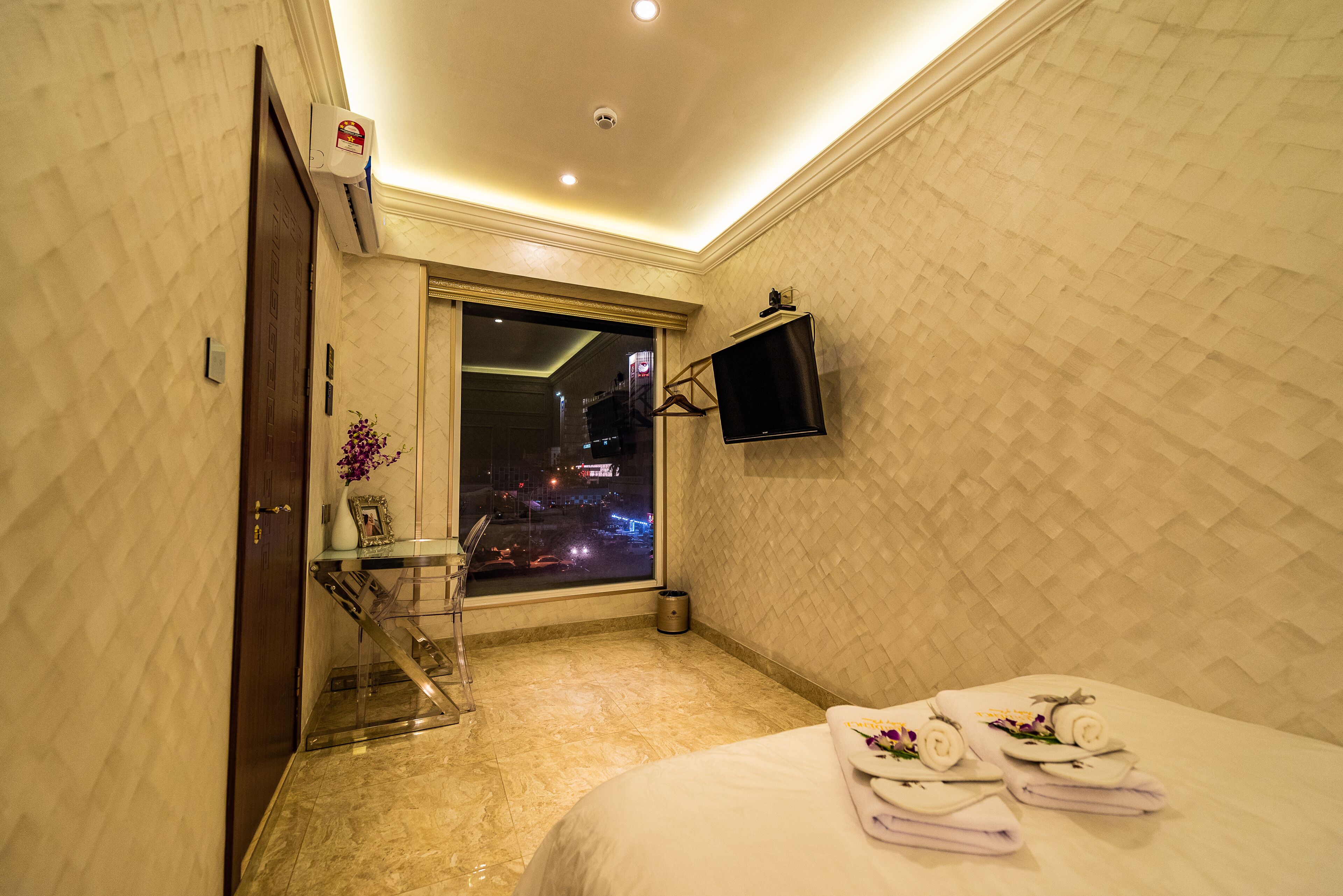 A hotel room with a bed, a TV mounted on the wall, a window with a city view, and a bathroom with a mirror and towel rack.