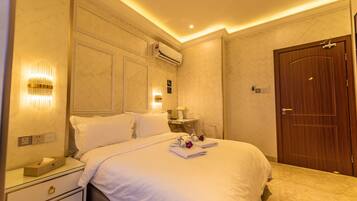 A hotel room with a neatly made bed, an air conditioning unit, a bedside table with a lamp, and a wooden door.