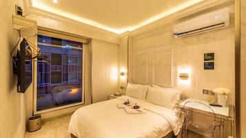 A hotel room with a bed, a desk with a chair, a TV mounted on the wall, and a window with a view of buildings.