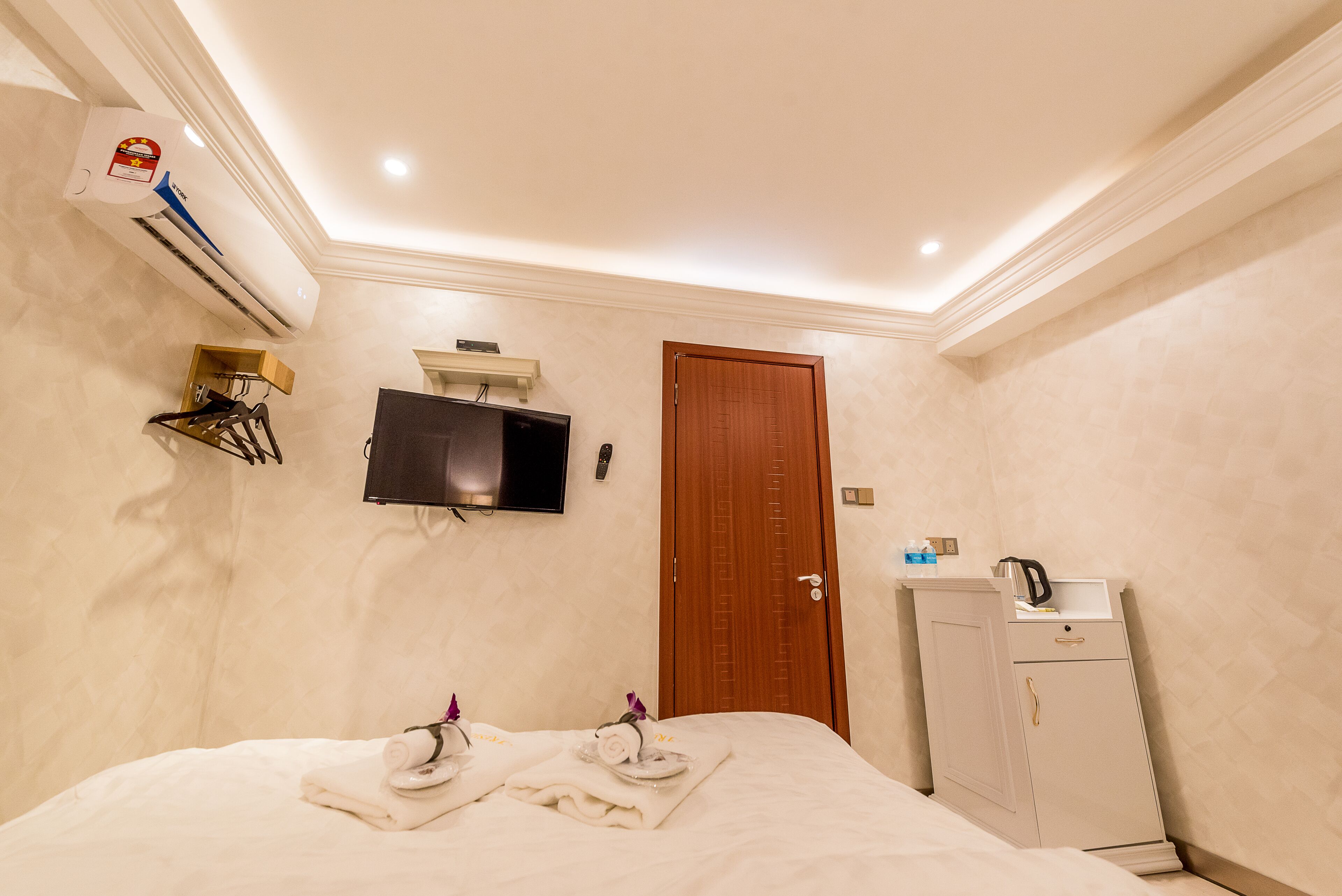 A hotel room with a bed, a TV mounted on the wall, an air conditioning unit, a wooden door, and a small cabinet.