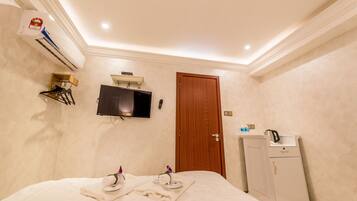 A hotel room with a bed, a TV mounted on the wall, an air conditioning unit, a wooden door, and a small cabinet.