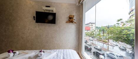 A hotel room with a bed, a TV mounted on the wall, and a view of the street through a large window.