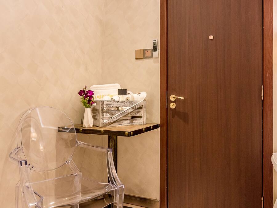 A hotel room with a wooden door, a clear plastic chair, a small table with toiletries, and a vase with flowers.