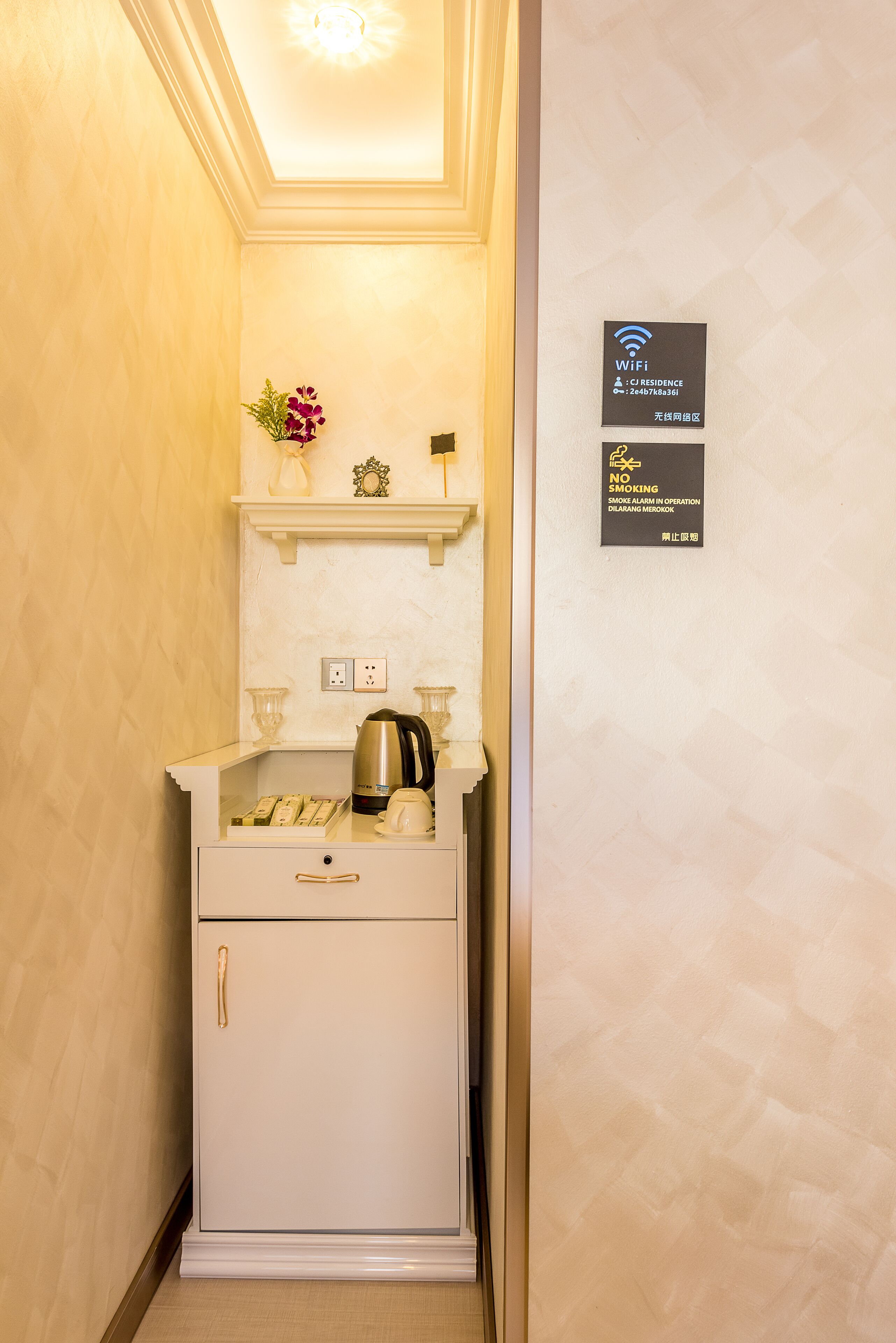 A compact kitchenette with a coffee maker, a kettle, and a small shelf with decorative items. There are signs indicating WiFi availability and a no-smoking policy.