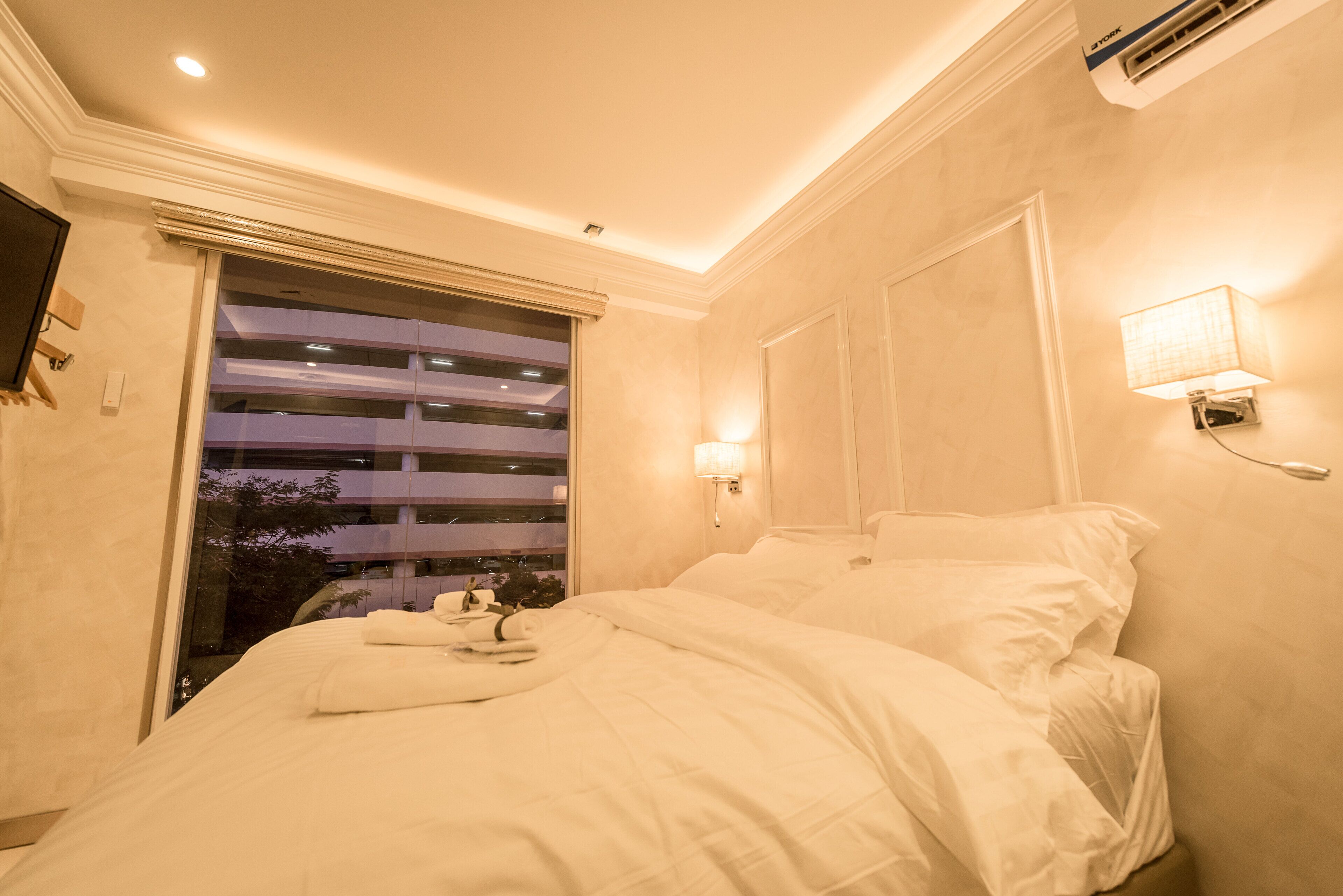 A hotel room with a bed, a window with blinds, a wall-mounted lamp, and an air conditioning unit.