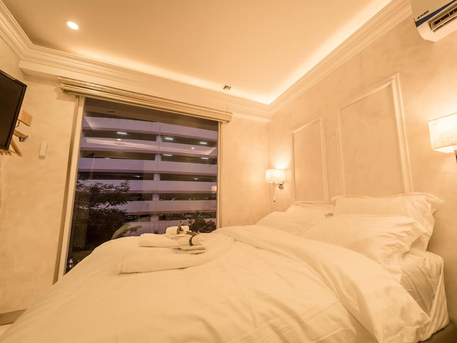 Une chambre d’hôtel avec un lit, une fenêtre avec des stores, une lampe fixée au mur et un appareil de climatisation.