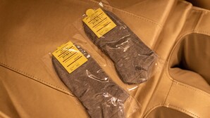 Zwei Paare grauer Socken in Plastikverpackung auf einer hellbraunen Lederoberfläche.