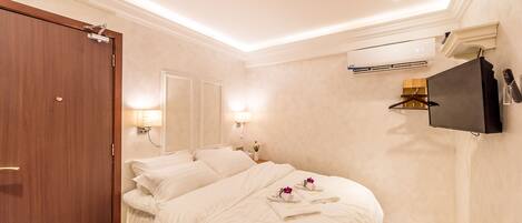 A hotel room with a bed, a wooden door, a wall-mounted TV, and an air conditioning unit.