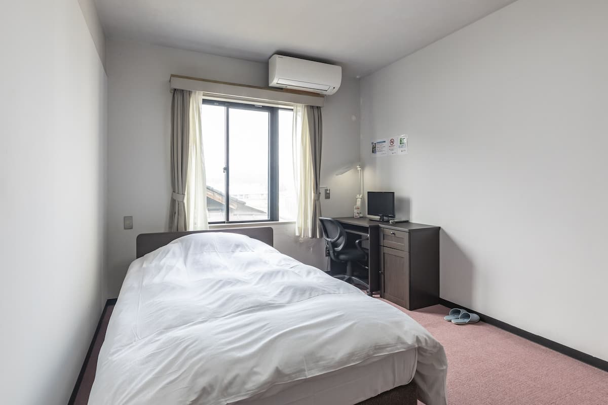 Une chambre d’hôtel équipée d’un lit, d’un bureau avec un ordinateur, d’un climatiseur et d’une fenêtre avec des rideaux.