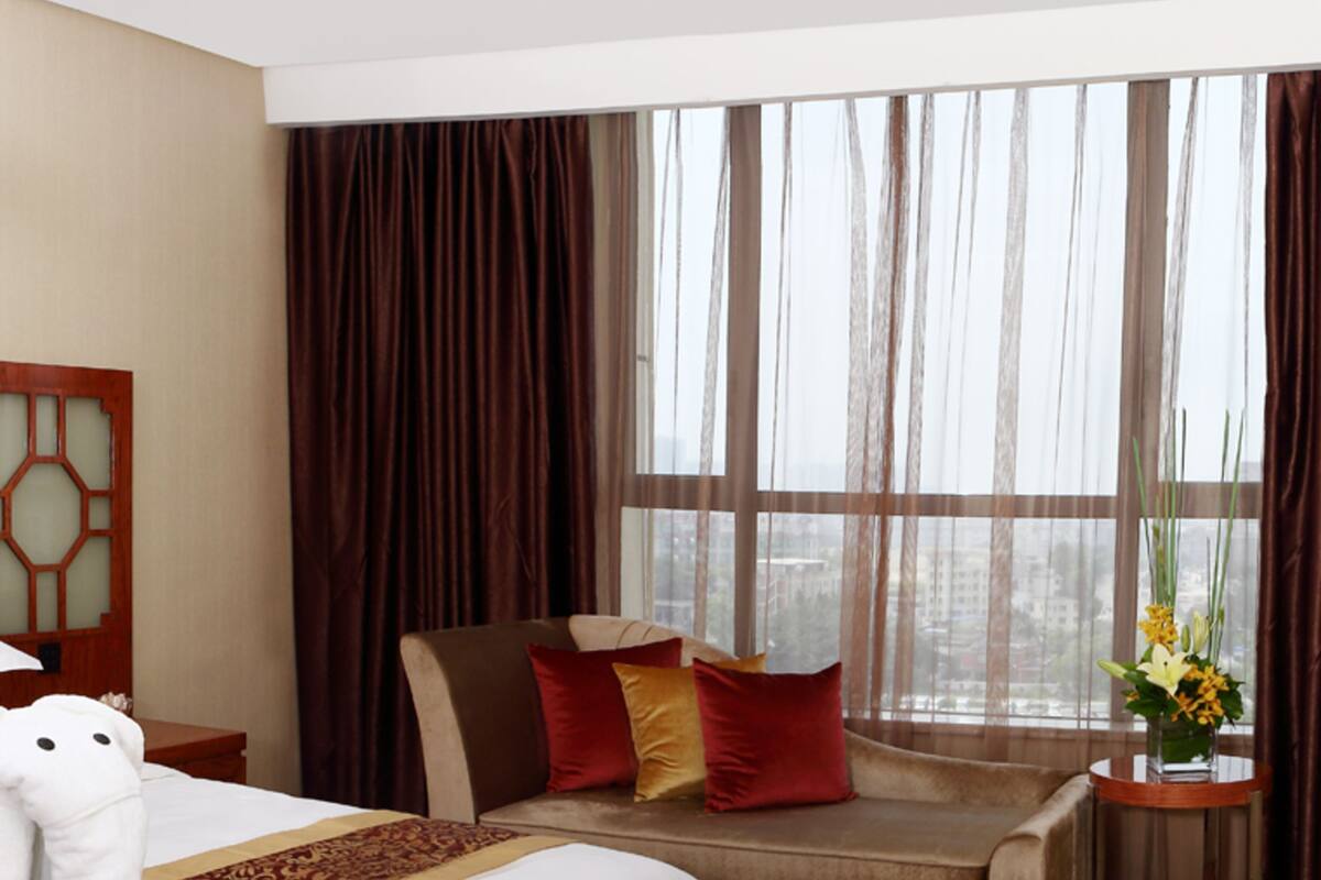Habitación de hotel con una cama grande, un sofá, una mesita con un florero y vistas a la ciudad a través de amplias ventanas.
