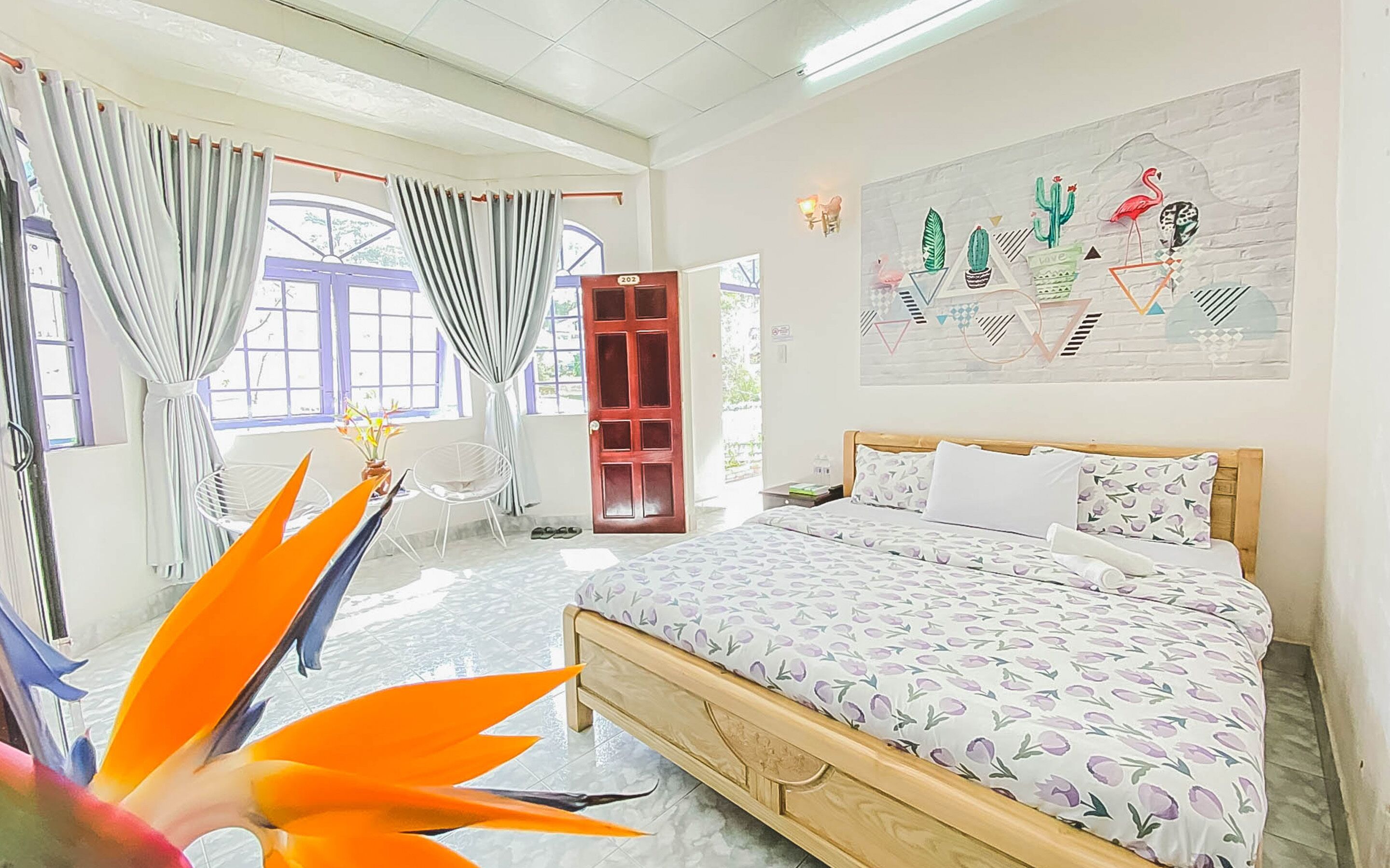 Ein Schlafzimmer mit Bett, einem Fenster mit Vorhängen, einer Wand mit Kakteen- und Flamingodekoration und einer Pflanze im Vordergrund.