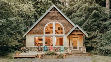 Una cabaña de madera con cimientos de piedra, ventanas amplias y un porche con sillas Adirondack.