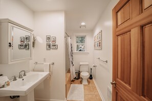 A bathroom with a white sink, toilet, and shower. There are framed pictures on the walls and a wooden door. - Mt Baker Lodging's Cabin #1 - With Wifi, Hot Tub, Pet Friendly (Deming)