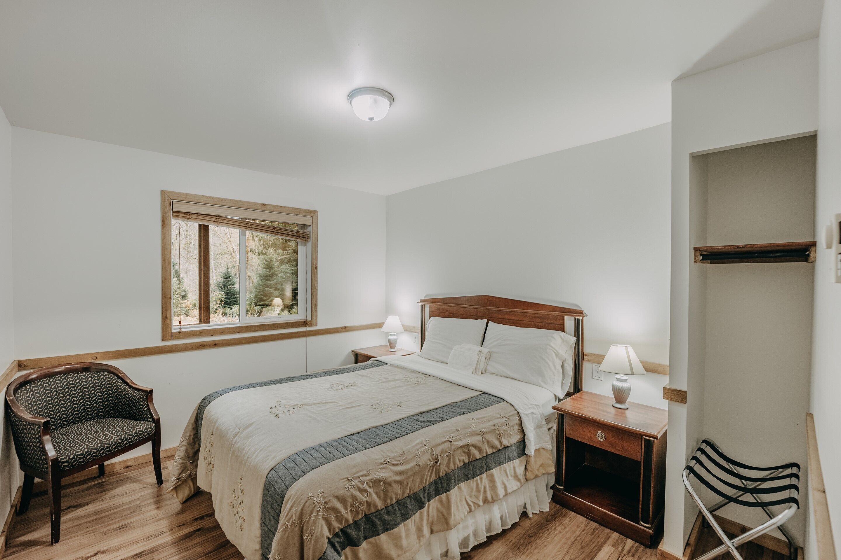 Un dormitorio con una cama grande, mesitas de noche de madera, una silla, una ventana con vista a árboles y una lámpara en el techo.