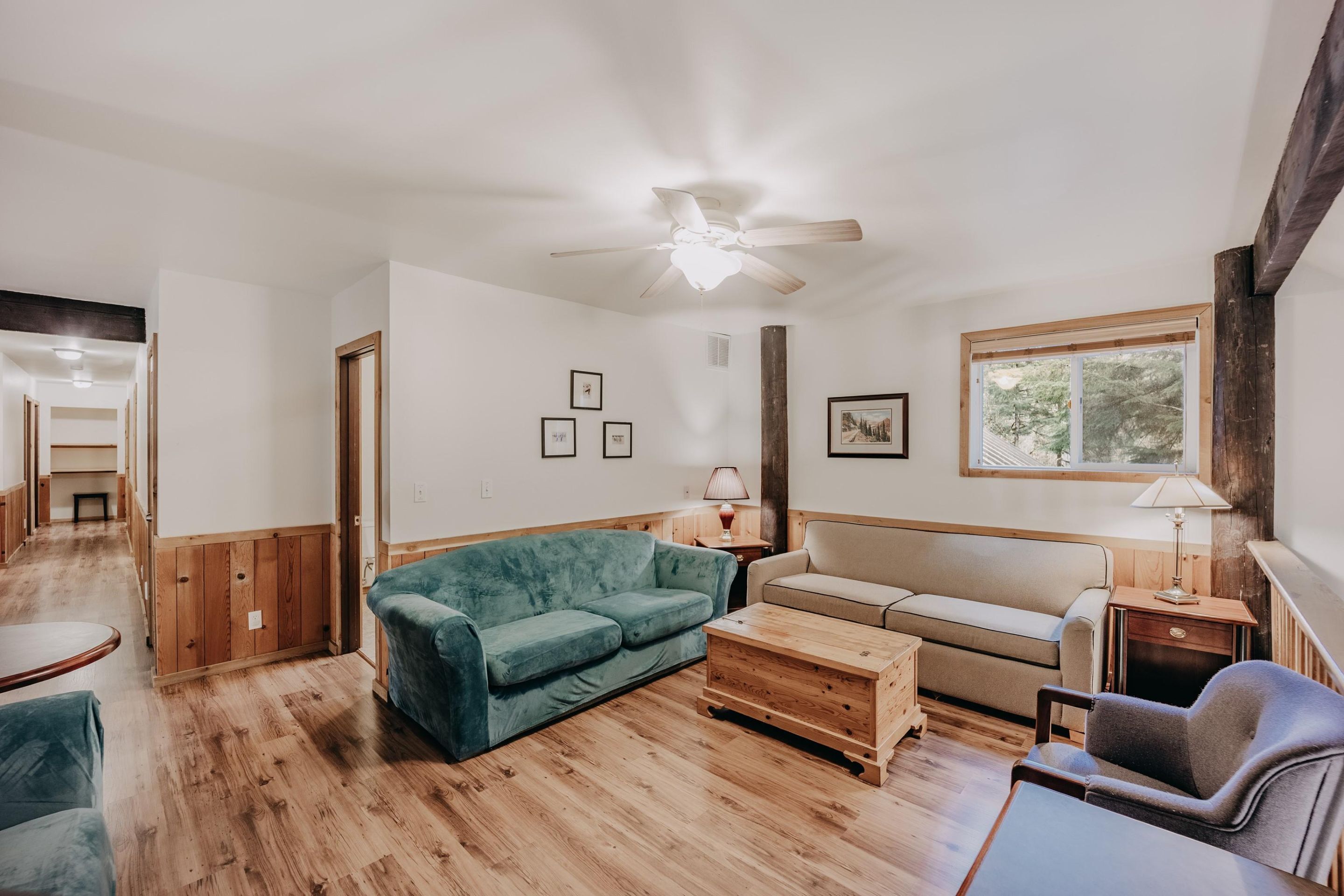Amplia sala de estar con piso de madera, ventilador de techo, un sofá verde, un sofá color beige, una mesa de centro de madera y un sillón gris.