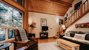 A cozy living room with a wooden ceiling, a large window, a leather chair, a sofa with a 'RELAX' pillow, a wooden bench, and a small table. - Snowline Cabin #4 - Pet-friendly Cedar Cabin With a Private Outdoor hot Tub! (Deming)