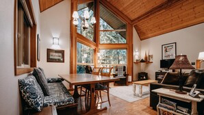 A cozy living room with a wooden ceiling, large windows, a dining table, and a sofa. - Snowline Cabin #4 - Pet-friendly Cedar Cabin With a Private Outdoor hot Tub! (Deming)