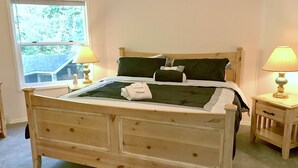 Ein hölzernes Bett mit Kopfteil, zwei Nachttische mit Lampen, ein Fenster mit Blick auf Bäume und ein Teppichboden.