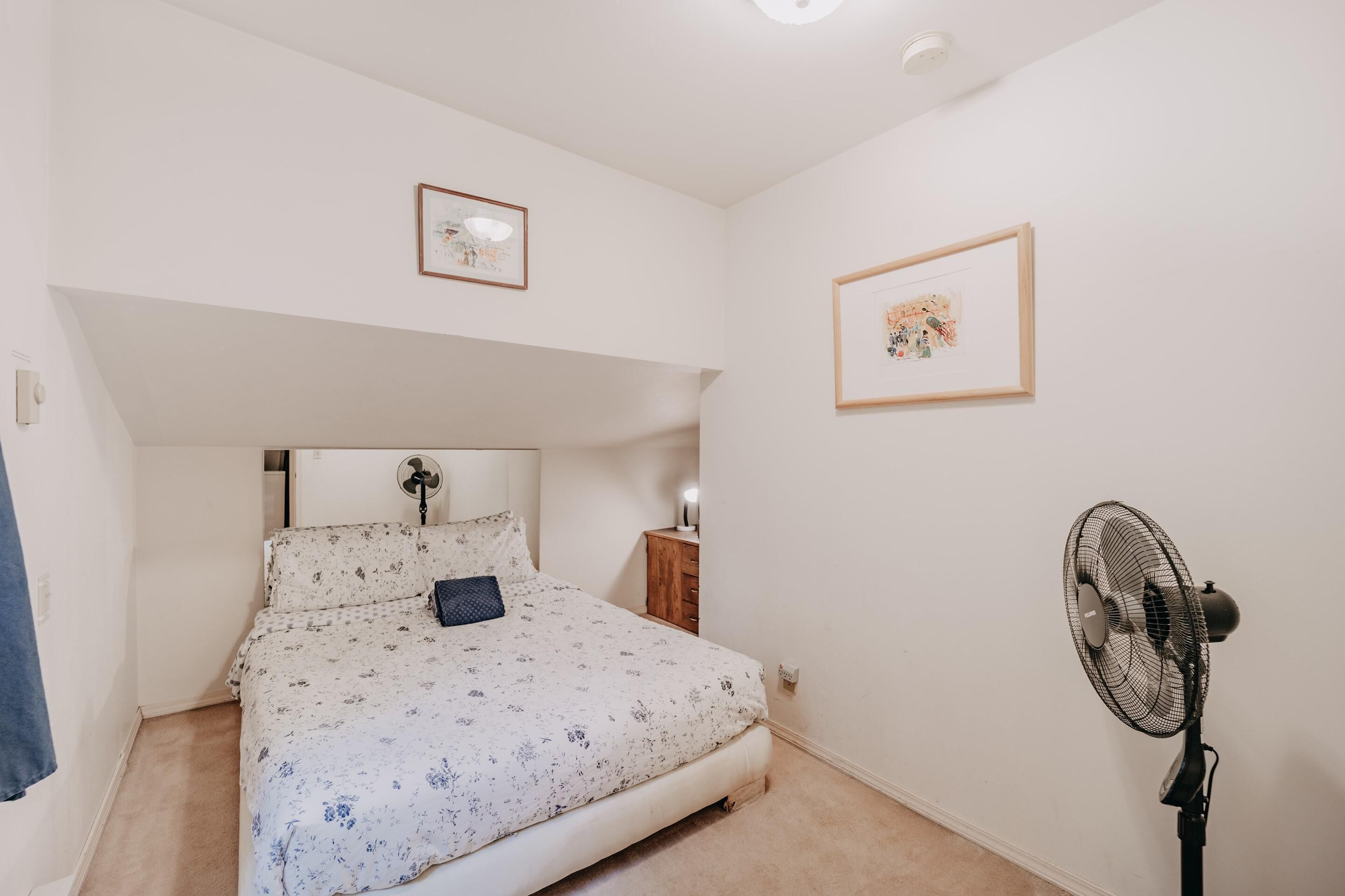 A bedroom with a bed, a standing fan, a nightstand, and two framed pictures on the wall.