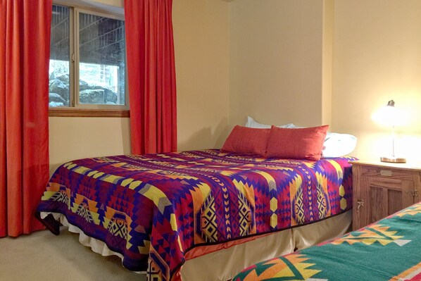 A bedroom with a bed covered in a colorful quilt, a wooden nightstand with a lamp, and a window with red curtains. - Snowline Cabin #29 - An Ultra Custom Family Vacation Home! (Deming)