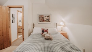 Een slaapkamer met een geruite plaid, een ingelijst kunstwerk aan de muur, een bedlamp en een houten deur die naar een andere kamer leidt.