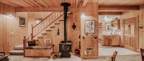 A cozy wooden cabin interior with a central wood-burning stove, a staircase, and a kitchen area.