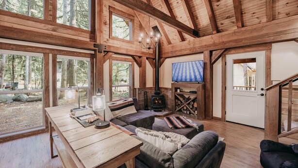 A cozy cabin interior with a large wooden dining table, comfortable seating, a fireplace, and large windows offering a view of the forest. - Glacier Springs Cabin #39 - New Modern-rustic Cabin With Air Conditioning! (Deming)