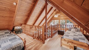 A wooden attic bedroom with two beds, a balcony, and a view of the outside.