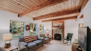 A cozy living room with a fireplace, a sofa, a television, and large windows offering a view of the outdoors. - Mt Baker Lodging - Cabin 67 - Private 2-story Cabin With a Private hot tub (Maple Falls)