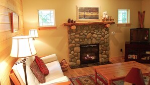 A cozy living room with a stone fireplace, a white sofa, a wooden coffee table, and a television on a wooden stand. - Glacier Springs Cabin 2bedrooms, 2 Baths - hot tub & Pet Friendly! (Deming)
