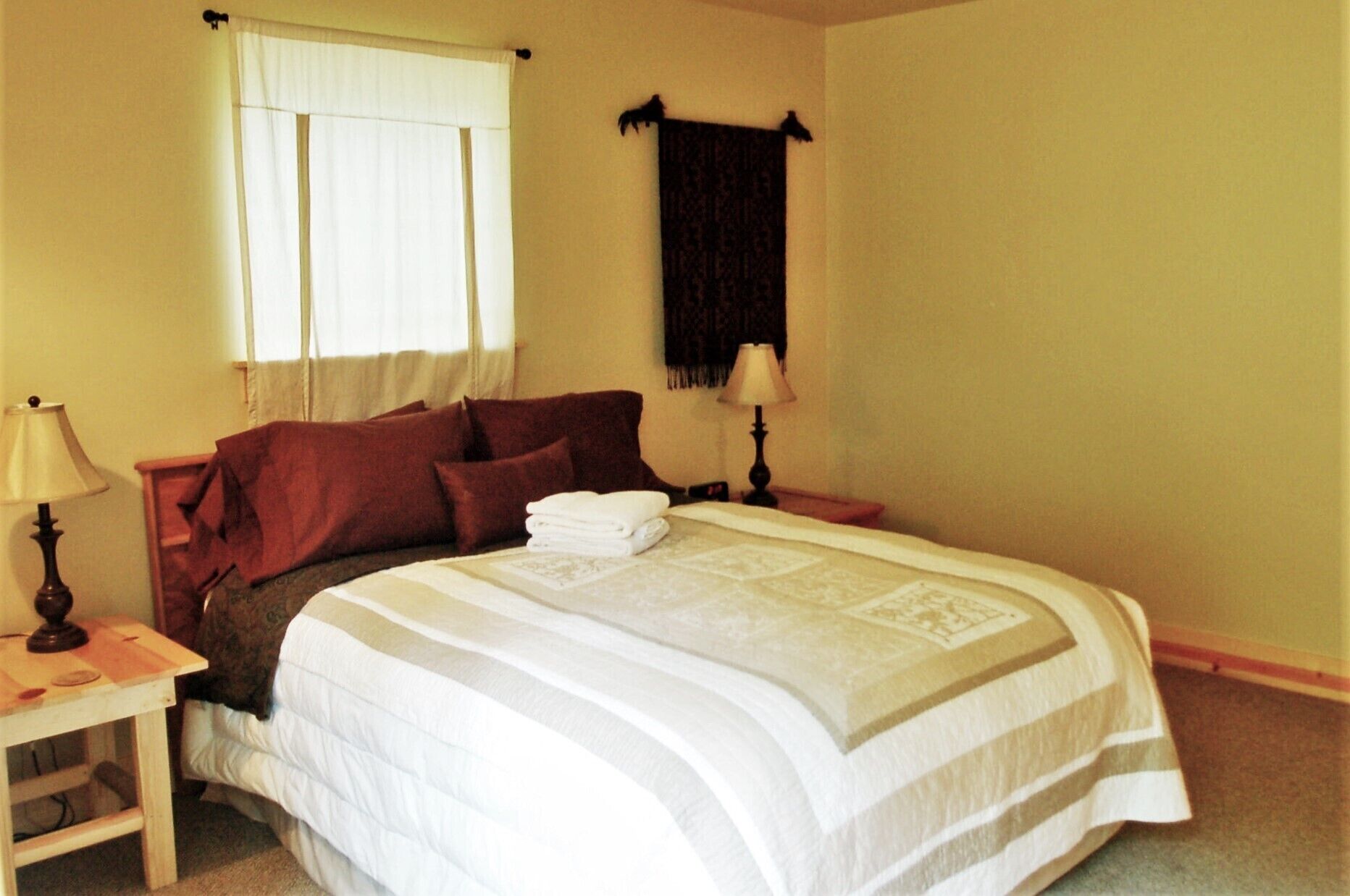 A neatly made bed with a white and gray striped bedspread, a wooden nightstand with a lamp, and a window with white curtains.