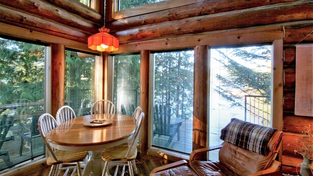 A wooden dining area with a round table and chairs, large windows with blinds, and a cozy seating area with a cushioned chair. - Cabin #97 - 'pinecone' Log Cabin at the Lake That is Pet-friendly! (Deming)