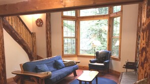 A cozy living room with a wooden staircase, a blue sofa, a patterned armchair, a coffee table, and a view of greenery through large windows. - Snowline Cabin #25 A Country-style pet Friendly Cabin With hot tub and Wifi! (Deming)