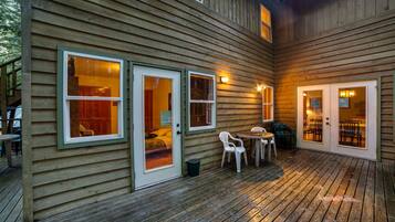 A wooden cabin with a deck, featuring a dining area with chairs and a table, and two doors leading to interior rooms.