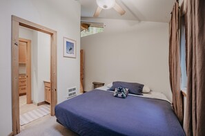 Une chambre à coucher comprenant un lit, un ventilateur de plafond, une fenêtre avec des rideaux, une porte donnant sur une salle de bain et une petite table de chevet.