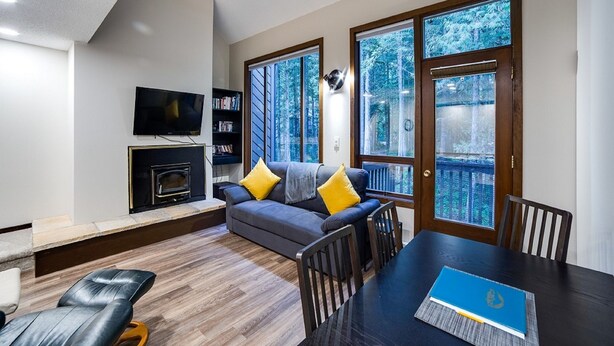 A modern living room with a fireplace, a sofa, a dining table, and large windows offering a view of trees. - Snowater Condo #story Condo - Sleeps 4 - Close to Community Amenities! (Deming)