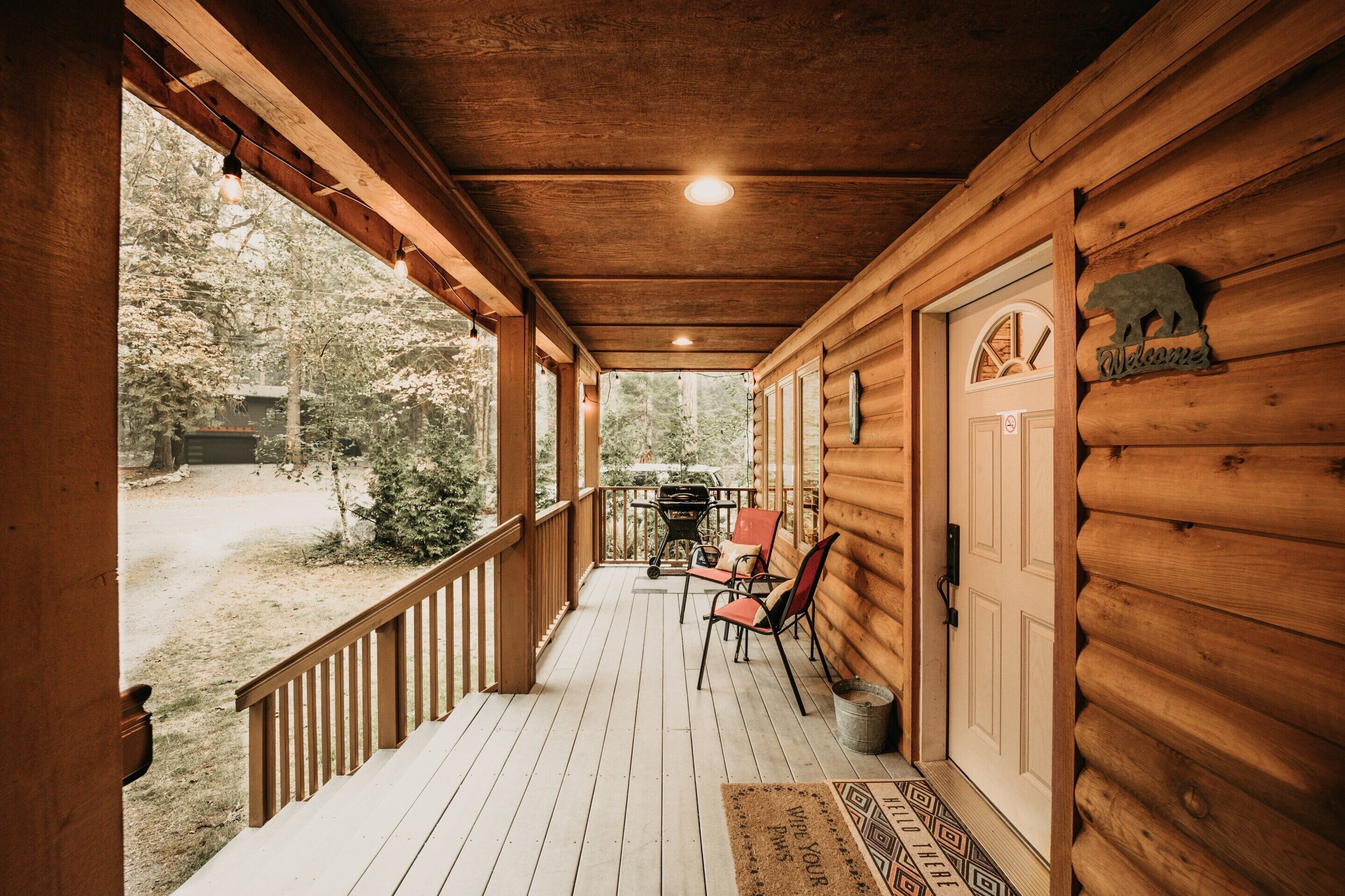 A wooden porch with a view of the surrounding forest, a barbecue grill, and a welcome sign.
