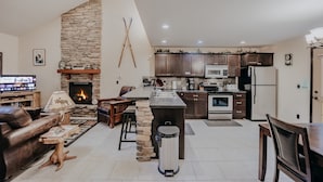 A modern living room with a stone fireplace, a kitchen with dark wooden cabinets, and a dining area.
