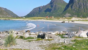 Paesaggio costiero con spiaggia rocciosa, un piccolo borgo con case a tetti rossi e una catena montuosa sullo sfondo.
