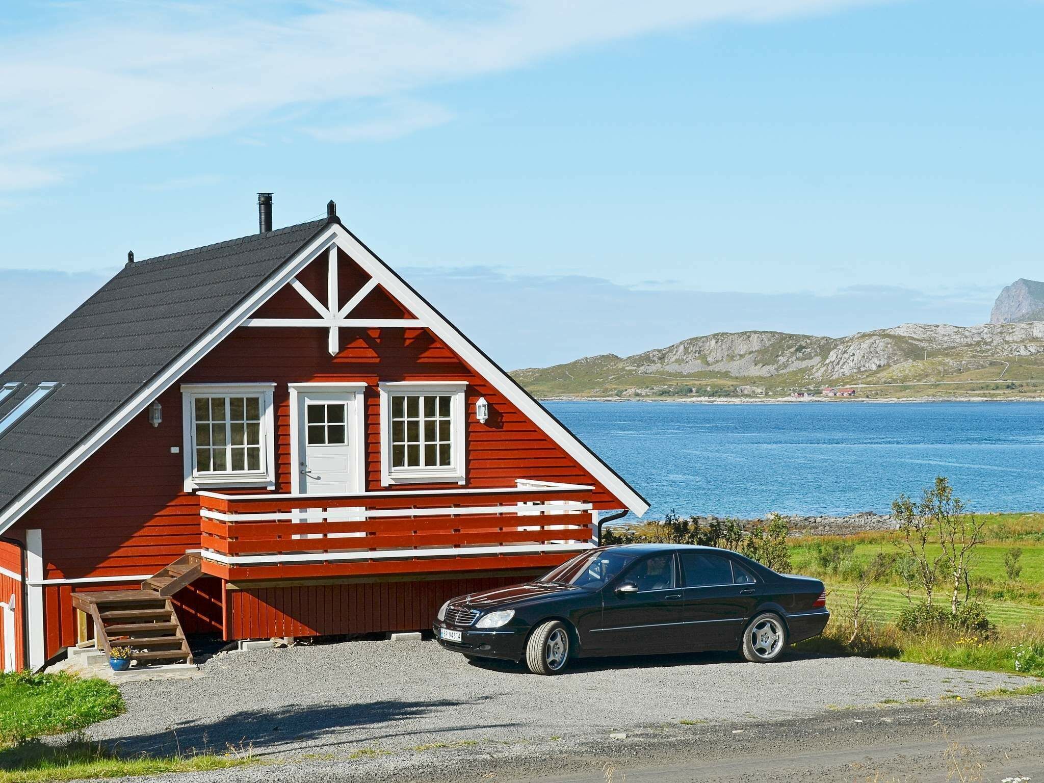 A red house with a car parked in front, near a body of water and hills.