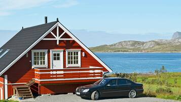 A red house with a car parked in front, near a body of water and hills.