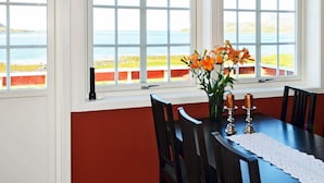Zona pranzo con tavolo apparecchiato per quattro persone, un vaso con fiori arancioni e vista sul mare attraverso ampie finestre.