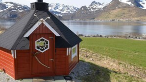 Un piccolo chalet di legno rosso con tetto nero, un camino e una finestra esagonale, affacciato su un lago con montagne sullo sfondo.
