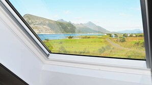 Paesaggio montuoso con un lago, prati verdi e alcuni edifici, vista da una finestra.